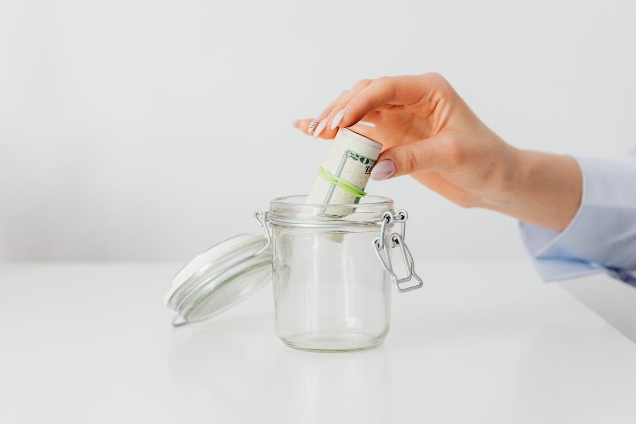 A close-up of a womans hand putting rolled US dollar bills into a glass jar, symbolizing saving and budgeting.